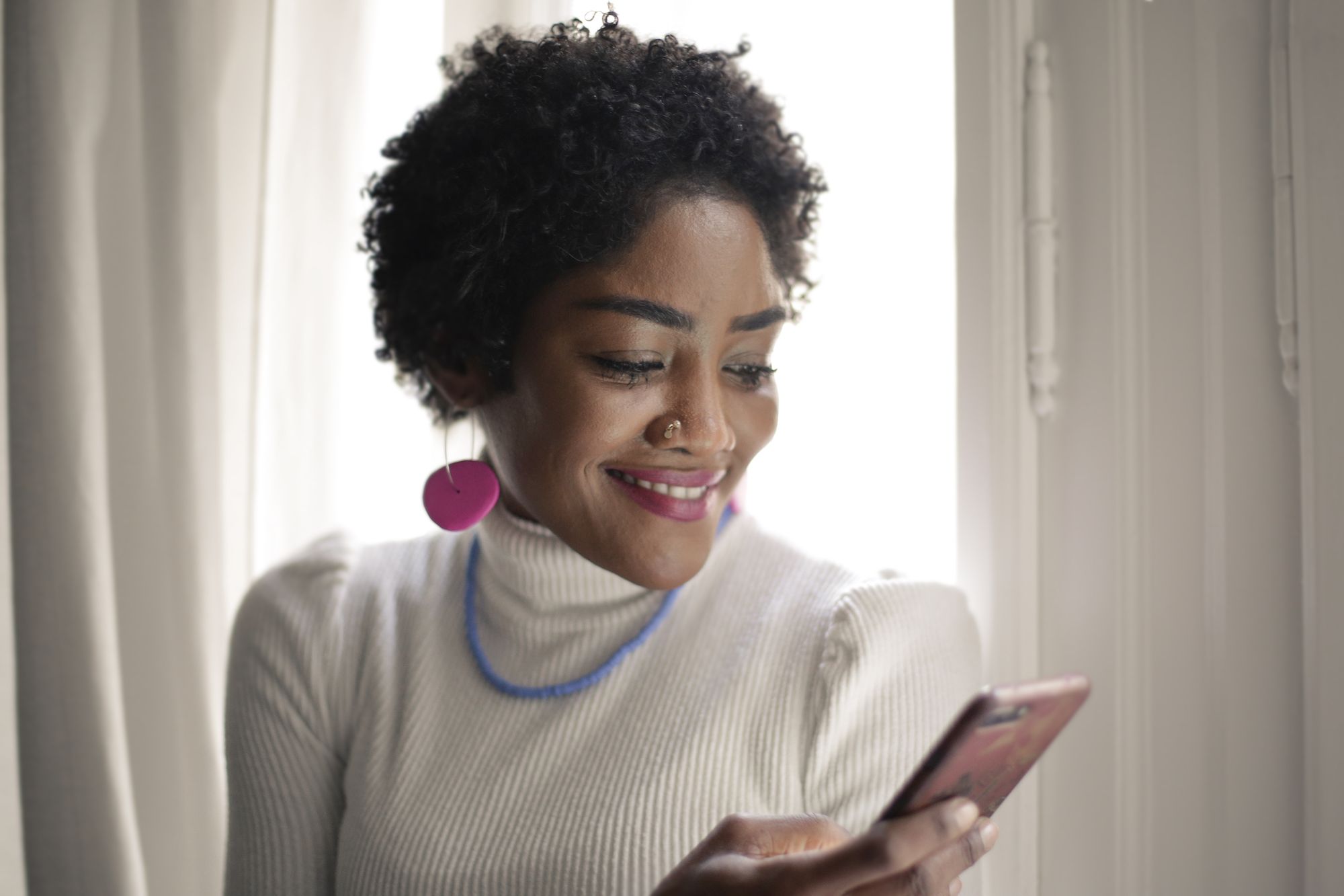 A woman smiling at her phone by a window